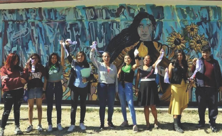 Students standing in front of a mural raising up one hand.